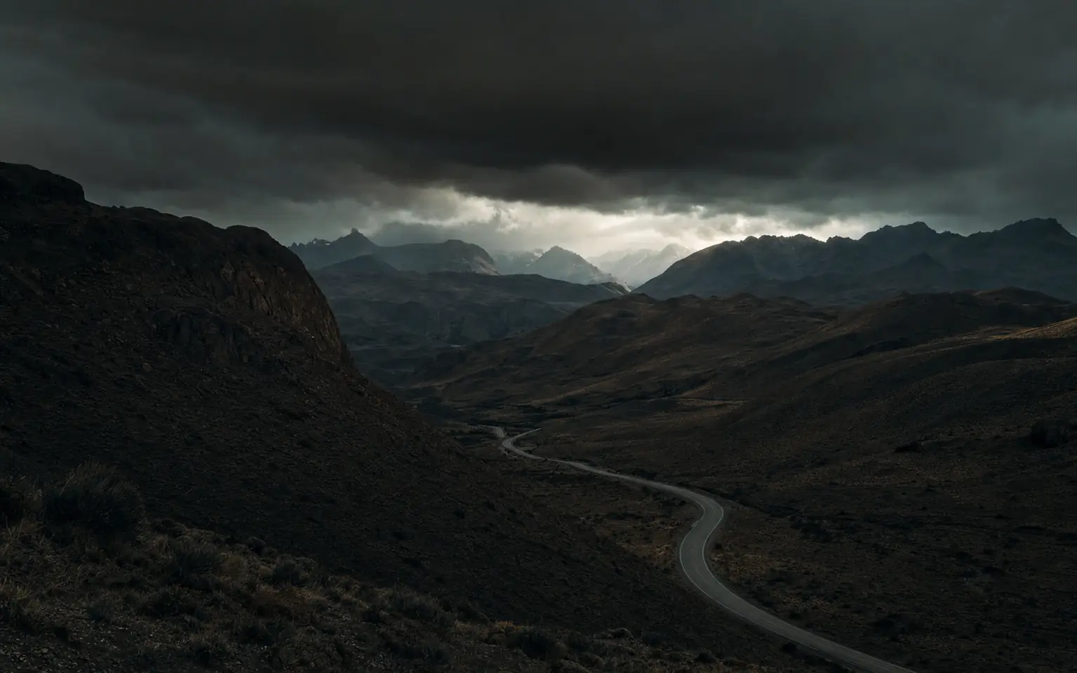An empty two-lane highway winding through vast arid Andean mountain ranges under a storm sky
