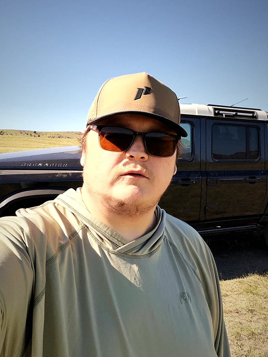 Taylor Guglielmino in a cap and sunglasses on the Montana high plains, his INEOS Grenadier Quartermaster parked on the two-track behind him