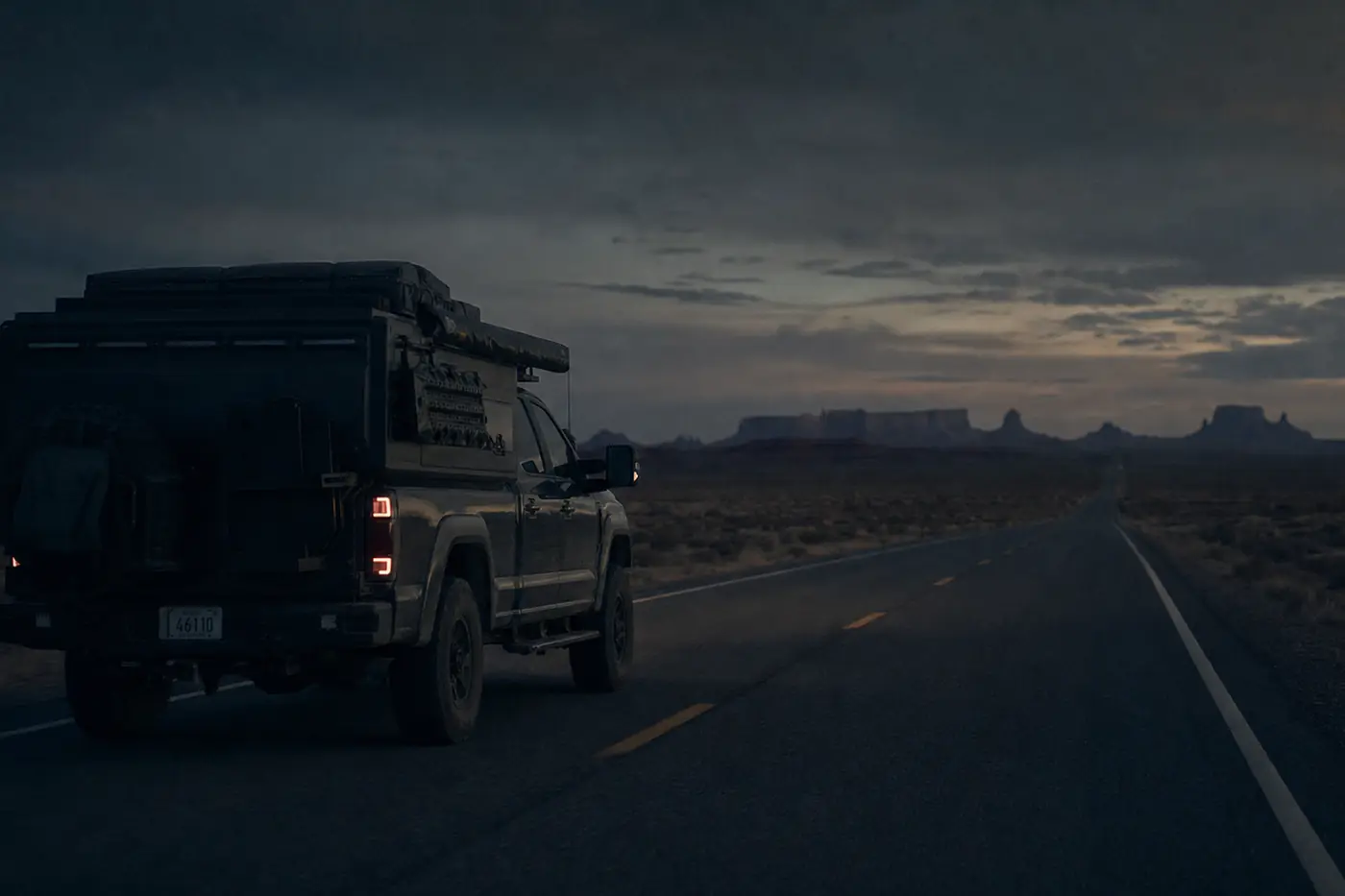 A full-size overland pickup truck with a bed camper travelling an empty desert highway at dusk