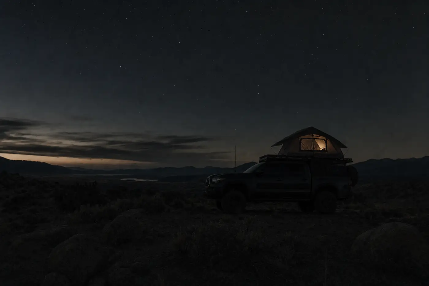 A mid-size overland pickup truck with a rooftop tent parked at a remote high desert camp at deep twilight